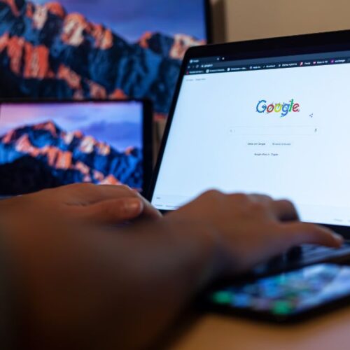 Person nutzt die Google-Suchmaschine auf einem Laptop, daneben ein Smartphone und ein weiterer Bildschirm im Hintergrund.
