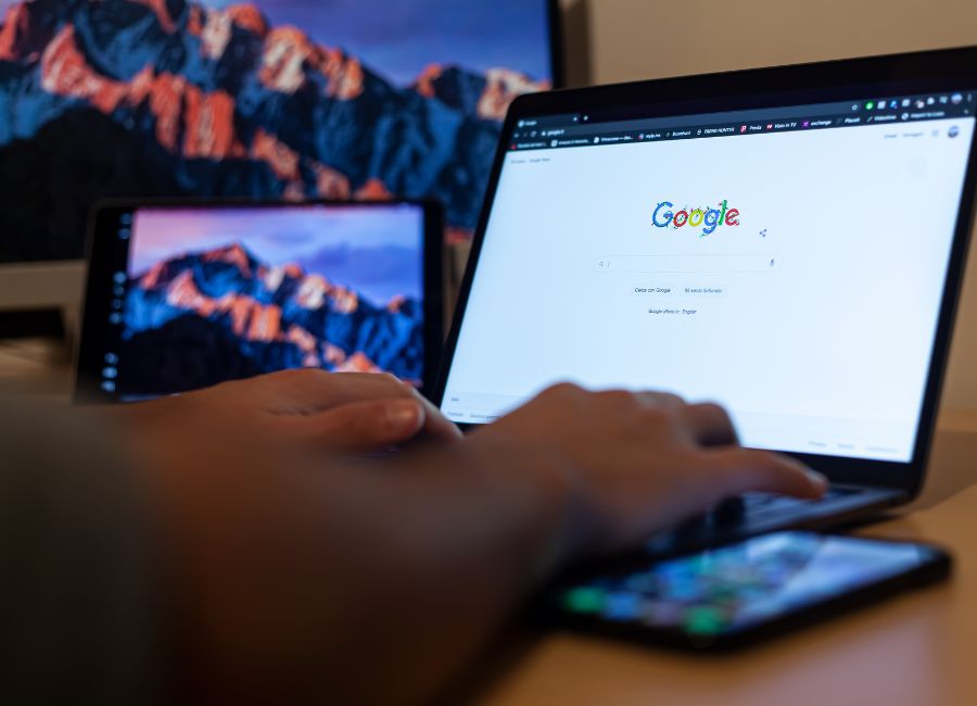 Person nutzt die Google-Suchmaschine auf einem Laptop, daneben ein Smartphone und ein weiterer Bildschirm im Hintergrund.