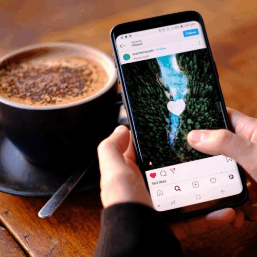 Eine Person scrolls am Handy durch Instagram und likes Bilder. Eine Tasse Kaffee im Hintergrund.